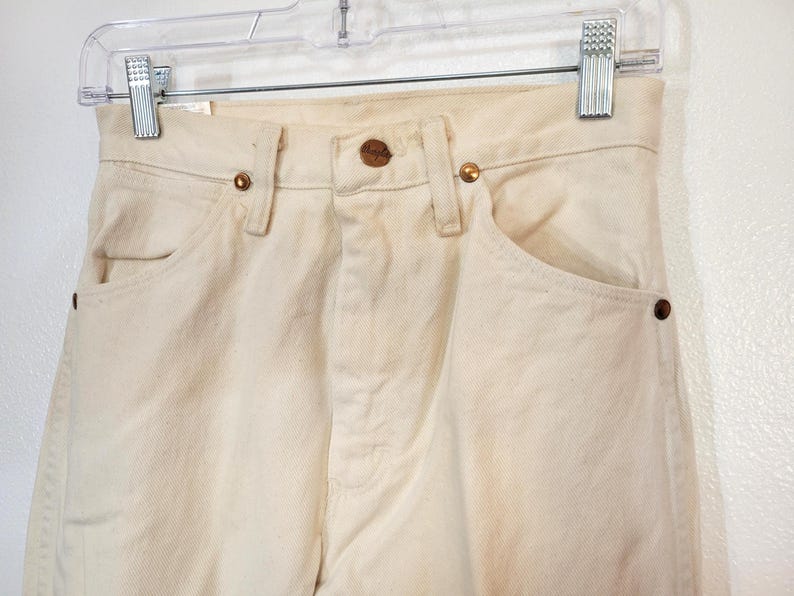 May include: Close-up of cream-colored denim jeans hanging on a clear hanger. The jeans have a classic five-pocket design with copper-colored rivets and a button closure. The fabric appears slightly worn.