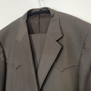 May include: A brown suit jacket with a notched lapel and a single button closure. The jacket has a subtle herringbone pattern and a pointed collar.
