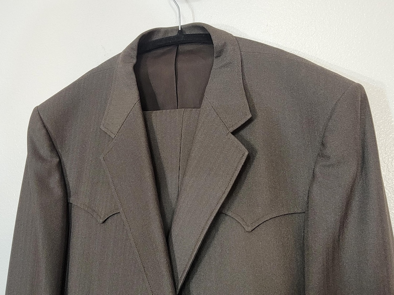 May include: A brown suit jacket with a notched lapel and a single button closure. The jacket has a subtle herringbone pattern and a pointed collar.