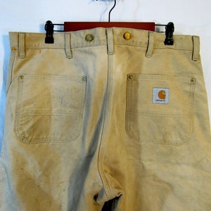 May include: A pair of tan work pants with two back pockets and a Carhartt logo patch. The pants have a worn look and are made of a durable fabric.