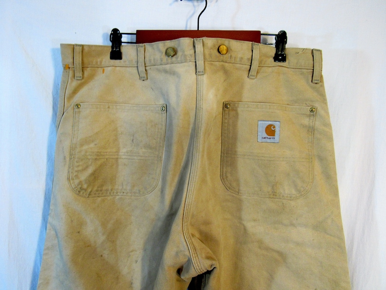 May include: A pair of tan work pants with two back pockets and a Carhartt logo patch. The pants have a worn look and are made of a durable fabric.
