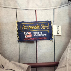 May include: Close-up of a light gray button-down shirt with vertical black and red stripes. The shirt features a label that reads "Panhandle Slim" with an American flag and the text "Proud to be American, Permanently Pressed."