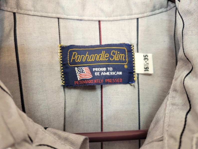 May include: Close-up of a light gray button-down shirt with vertical black and red stripes. The shirt features a label that reads "Panhandle Slim" with an American flag and the text "Proud to be American, Permanently Pressed."