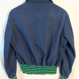 May include: Navy blue bomber jacket with a green and black striped elastic waistband and cuffs. The jacket has a collared neckline and is hanging on a wooden hanger. The back of the jacket is visible.