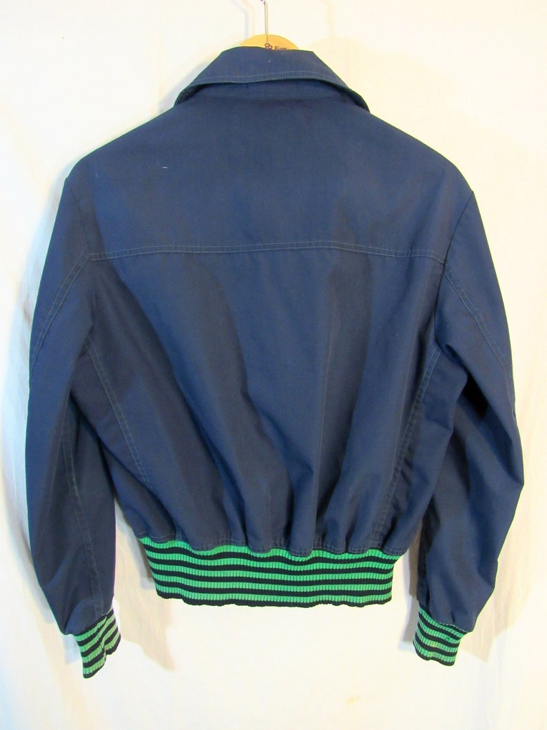 May include: Navy blue bomber jacket with a green and black striped elastic waistband and cuffs. The jacket has a collared neckline and is hanging on a wooden hanger. The back of the jacket is visible.