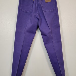 May include: Purple denim jeans with a classic five-pocket design. The jeans feature a brown leather patch and the Wrangler logo. The back pockets have the signature 'W' stitching. The jeans are hanging on a hanger.