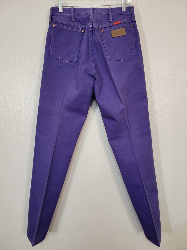 May include: Purple denim jeans with a classic five-pocket design. The jeans feature a brown leather patch and the Wrangler logo. The back pockets have the signature 'W' stitching. The jeans are hanging on a hanger.