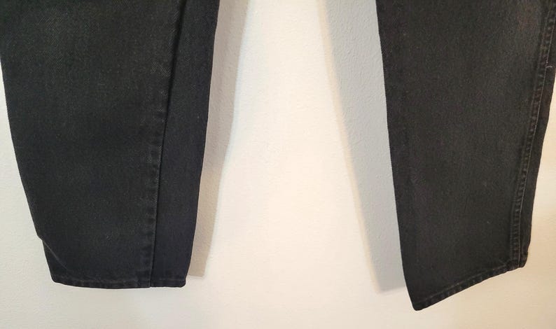 Puede incluir: Primer plano de unos vaqueros de mezclilla negros. Los vaqueros son de color negro liso y parecen estar hechos de un material de mezclilla resistente. La imagen muestra las perneras de los vaqueros, con las costuras y los dobladillos visibles.