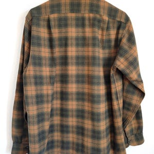 Pode incluir: Camisa de manga comprida com padr&atilde;o xadrez em tons de castanho e verde escuro. A camisa est&aacute; pendurada num cabide preto sobre um fundo branco. A parte de tr&aacute;s da camisa &eacute; vis&iacute;vel, com a gola e os punhos abotoados.