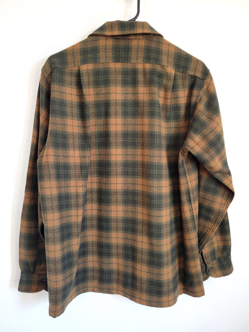 Pode incluir: Camisa de manga comprida com padr&atilde;o xadrez em tons de castanho e verde escuro. A camisa est&aacute; pendurada num cabide preto sobre um fundo branco. A parte de tr&aacute;s da camisa &eacute; vis&iacute;vel, com a gola e os punhos abotoados.