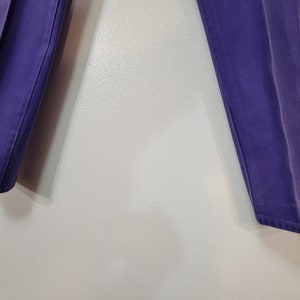 May include: A pair of purple pants. The pants are a solid color and appear to be made of a sturdy material. The legs are straight and the pants are hanging against a light-colored wall.