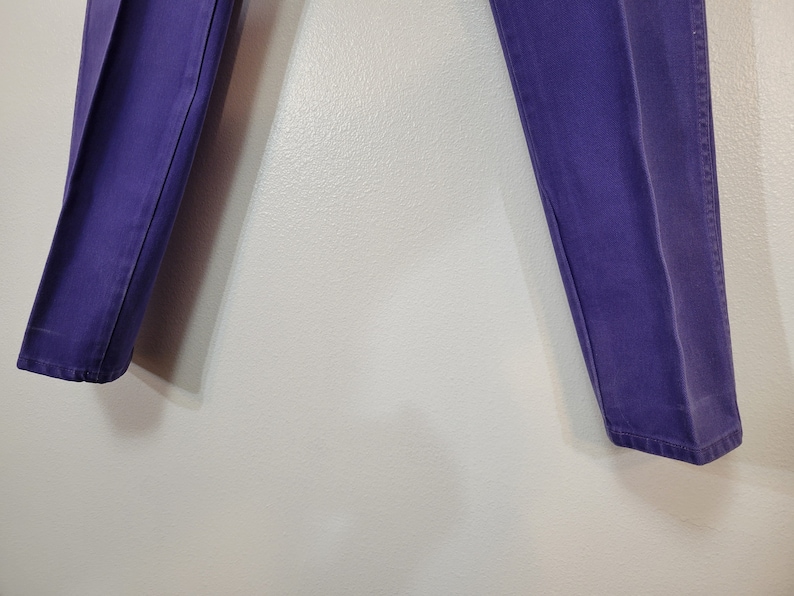 May include: A pair of purple pants. The pants are a solid color and appear to be made of a sturdy material. The legs are straight and the pants are hanging against a light-colored wall.