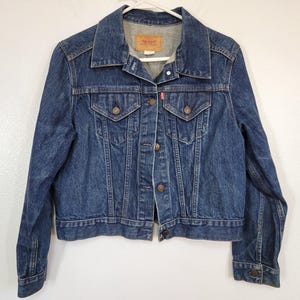 May include: A classic blue denim jacket with a button-up front, two chest pockets, and two side pockets. The jacket features a collar and long sleeves. A small red tag is visible on the chest pocket. The jacket is hanging on a white hanger.