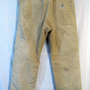 May include: A pair of beige work pants with a Carhartt logo patch on the back right pocket. The pants have a worn and faded look, suggesting they have been well-used.