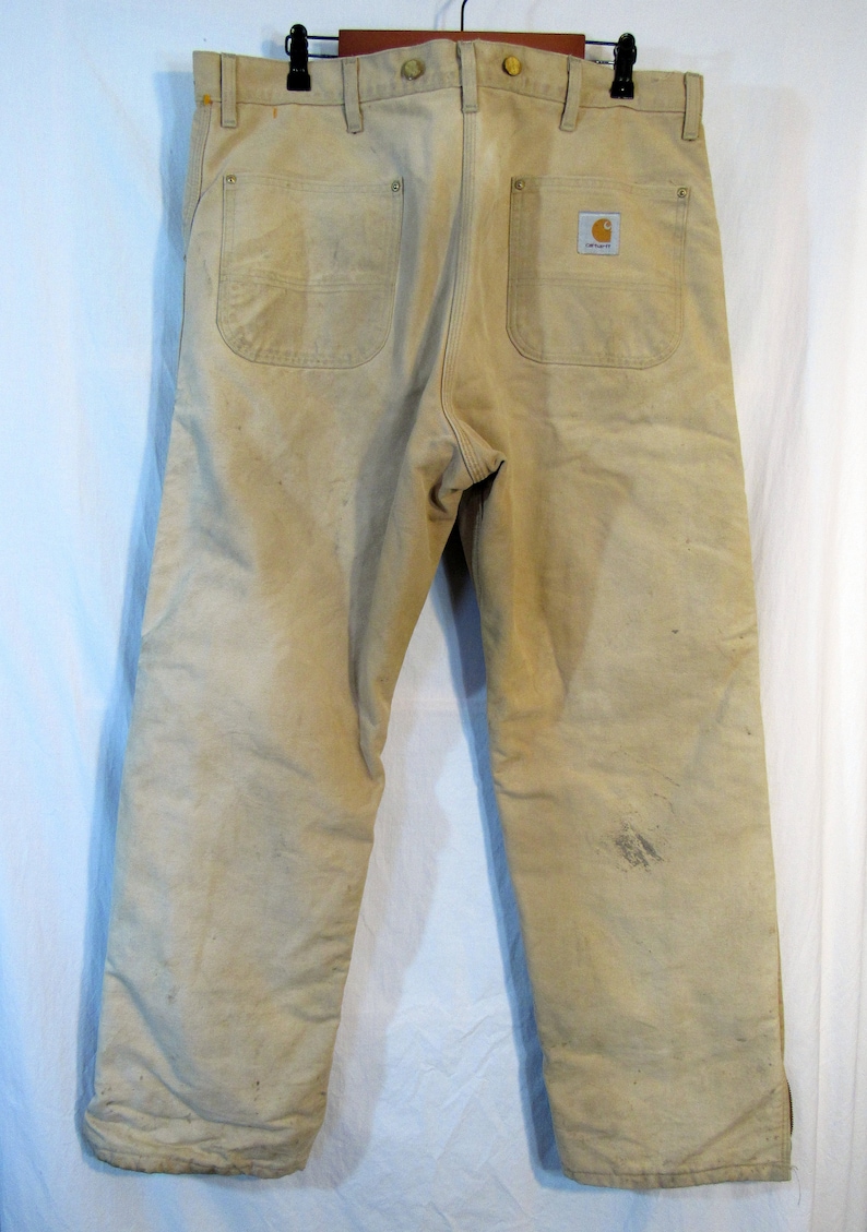 May include: A pair of beige work pants with a Carhartt logo patch on the back right pocket. The pants have a worn and faded look, suggesting they have been well-used.