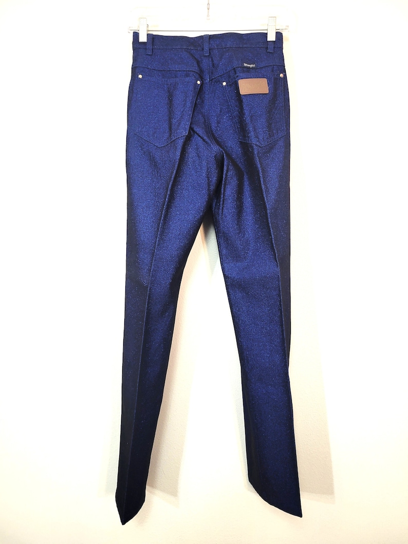 May include: Dark blue denim jeans with a classic five-pocket design. The jeans feature a straight leg cut and a brown leather patch on the back. The Wrangler logo is visible above the patch. The jeans are hanging on a white hanger.