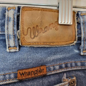 May include: Close-up of blue denim jeans with a brown leather patch featuring the brand name "Wrangler" in a Western-style font. The jeans have classic stitching and a small brown label with the brand name.