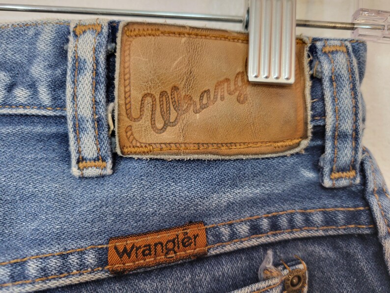 May include: Close-up of blue denim jeans with a brown leather patch featuring the brand name "Wrangler" in a Western-style font. The jeans have classic stitching and a small brown label with the brand name.
