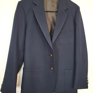May include: A navy blue blazer with two front pockets and gold-colored buttons. The blazer is hanging on a black hanger against a white background. The blazer has a classic, tailored design.