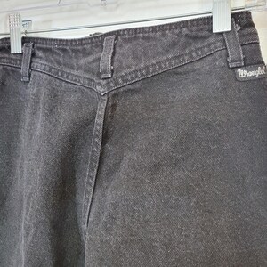 Puede incluir: Primer plano de unos vaqueros de mezclilla negros con cintura alta y un canes&uacute; en forma de V distintivo. Los vaqueros tienen trabillas y el logotipo de Wrangler. La tela parece ligeramente descolorida, lo que sugiere una est&eacute;tica vintage. Los vaqueros est&aacute;n colgados de una percha transparente.