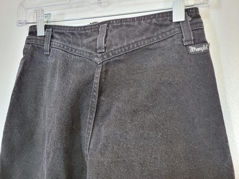 Puede incluir: Primer plano de unos vaqueros de mezclilla negros con cintura alta y un canes&uacute; en forma de V distintivo. Los vaqueros tienen trabillas y el logotipo de Wrangler. La tela parece ligeramente descolorida, lo que sugiere una est&eacute;tica vintage. Los vaqueros est&aacute;n colgados de una percha transparente.