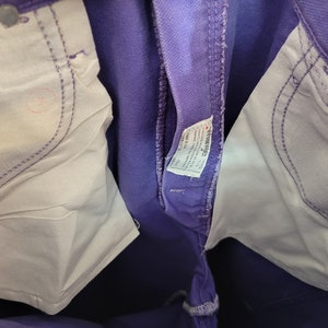 May include: Close-up of purple denim jeans with white pocket lining and silver rivets. The jeans have a classic five-pocket design and a visible brand tag. The stitching is a lighter purple color.