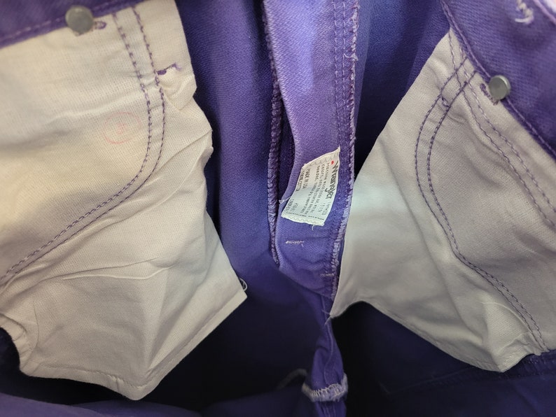 May include: Close-up of purple denim jeans with white pocket lining and silver rivets. The jeans have a classic five-pocket design and a visible brand tag. The stitching is a lighter purple color.