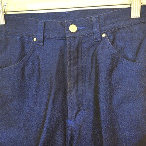 May include: Close-up of dark blue denim pants with a button and zipper closure. The pants have front and back pockets, belt loops, and gold-colored rivets. The pants are hanging on a metal hanger.