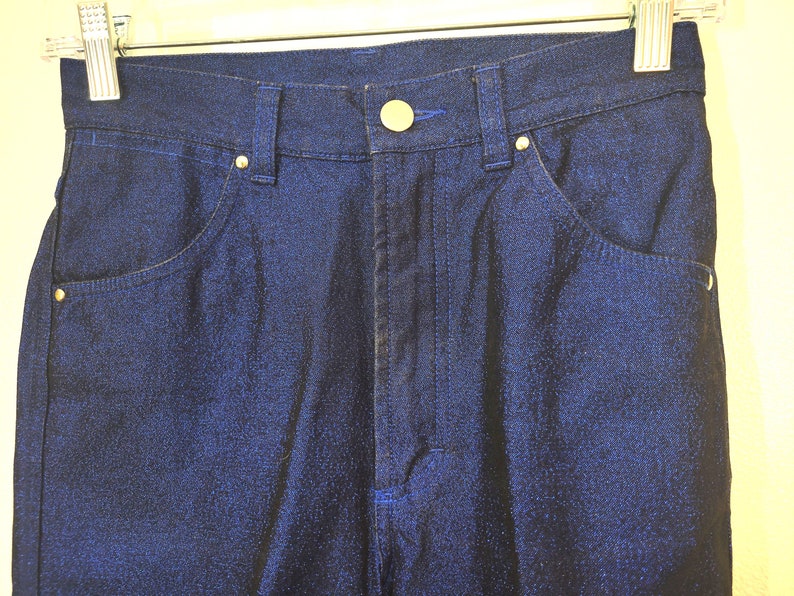May include: Close-up of dark blue denim pants with a button and zipper closure. The pants have front and back pockets, belt loops, and gold-colored rivets. The pants are hanging on a metal hanger.