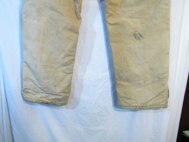 May include: A pair of off-white work pants with a worn and faded appearance. The pants have a straight leg and are made of a durable fabric.