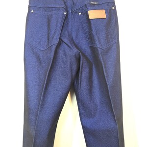 May include: Dark blue denim jeans with a classic five-pocket design. The jeans feature a leather patch with the brand name "Wrangler" on the back pocket. The jeans are hanging on a white hanger.