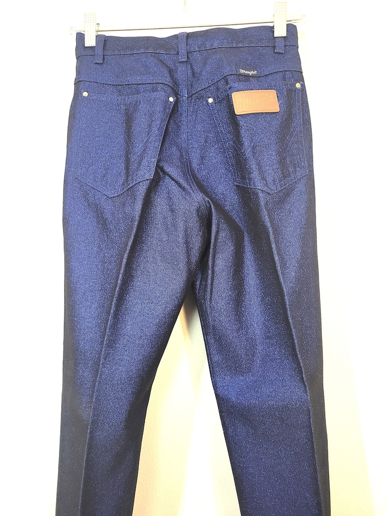 May include: Dark blue denim jeans with a classic five-pocket design. The jeans feature a leather patch with the brand name "Wrangler" on the back pocket. The jeans are hanging on a white hanger.