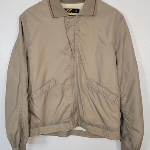 May include: A tan jacket with a collared neckline and a zipper closure. The jacket has two front pockets and ribbed cuffs and hem. The collar has a brown trim. The jacket is hanging on a wooden hanger.