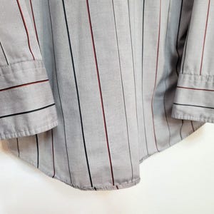 May include: Close-up of a gray button-down shirt with vertical stripes. The shirt features black and burgundy stripes, and the cuffs have matching stripes. The shirt is made of a lightweight fabric.