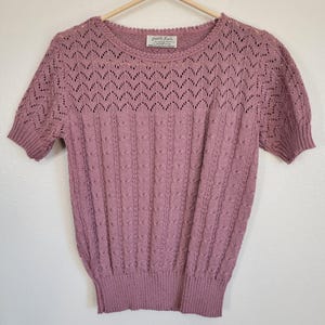 May include: A short-sleeved, mauve-colored knit top with a round neckline. The top features a textured pattern with openwork detailing across the chest and sleeves. The label reads "Fiddlesticks Knits". The garment is hanging on a wooden hanger.