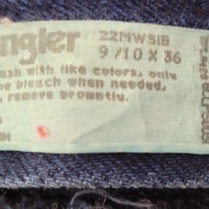 May include: Close-up of a light blue tag from Wrangler jeans. The tag includes the brand name, size information (9/10 x 36), and washing instructions. The tag is made in the USA and is 100% cotton.