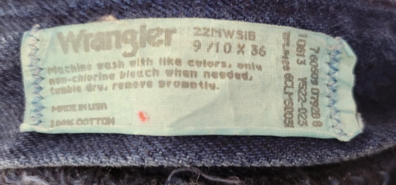 May include: Close-up of a light blue tag from Wrangler jeans. The tag includes the brand name, size information (9/10 x 36), and washing instructions. The tag is made in the USA and is 100% cotton.