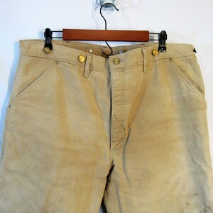 May include: A pair of tan work pants with brass buttons and a single front pocket. The pants are hanging on a wooden hanger.