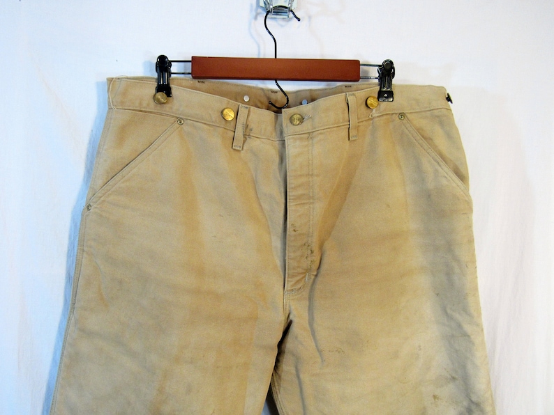 May include: A pair of tan work pants with brass buttons and a single front pocket. The pants are hanging on a wooden hanger.