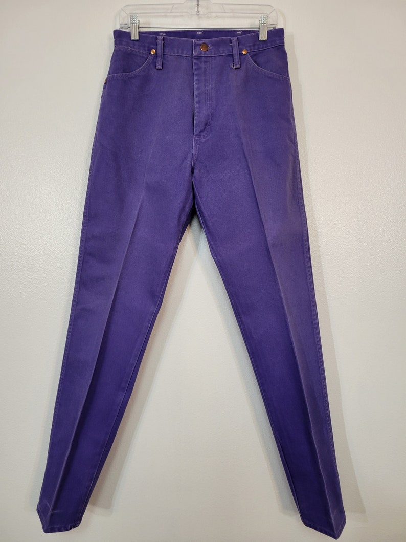 May include: A pair of purple denim pants, displayed on a hanger. The pants have a classic five-pocket design with a button and zipper closure. The denim is a solid, vibrant purple color.