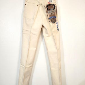 May include: Cream-colored Wrangler Cowboy Cut slim-fit jeans, size 14, hanging on a hanger. The jeans have a classic five-pocket design and a branded tag. The back pockets and seams are visible.