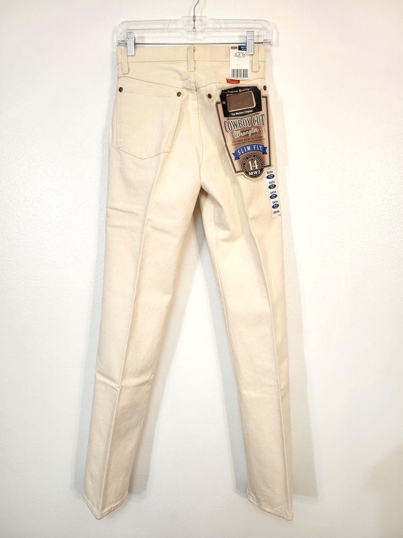 May include: Cream-colored Wrangler Cowboy Cut slim-fit jeans, size 14, hanging on a hanger. The jeans have a classic five-pocket design and a branded tag. The back pockets and seams are visible.
