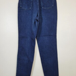 May include: Dark blue denim jeans with a classic five-pocket design. The jeans have a straight leg cut and a high waist. The Wrangler brand label is visible on the back pocket. The jeans are hanging on a hanger against a white background.