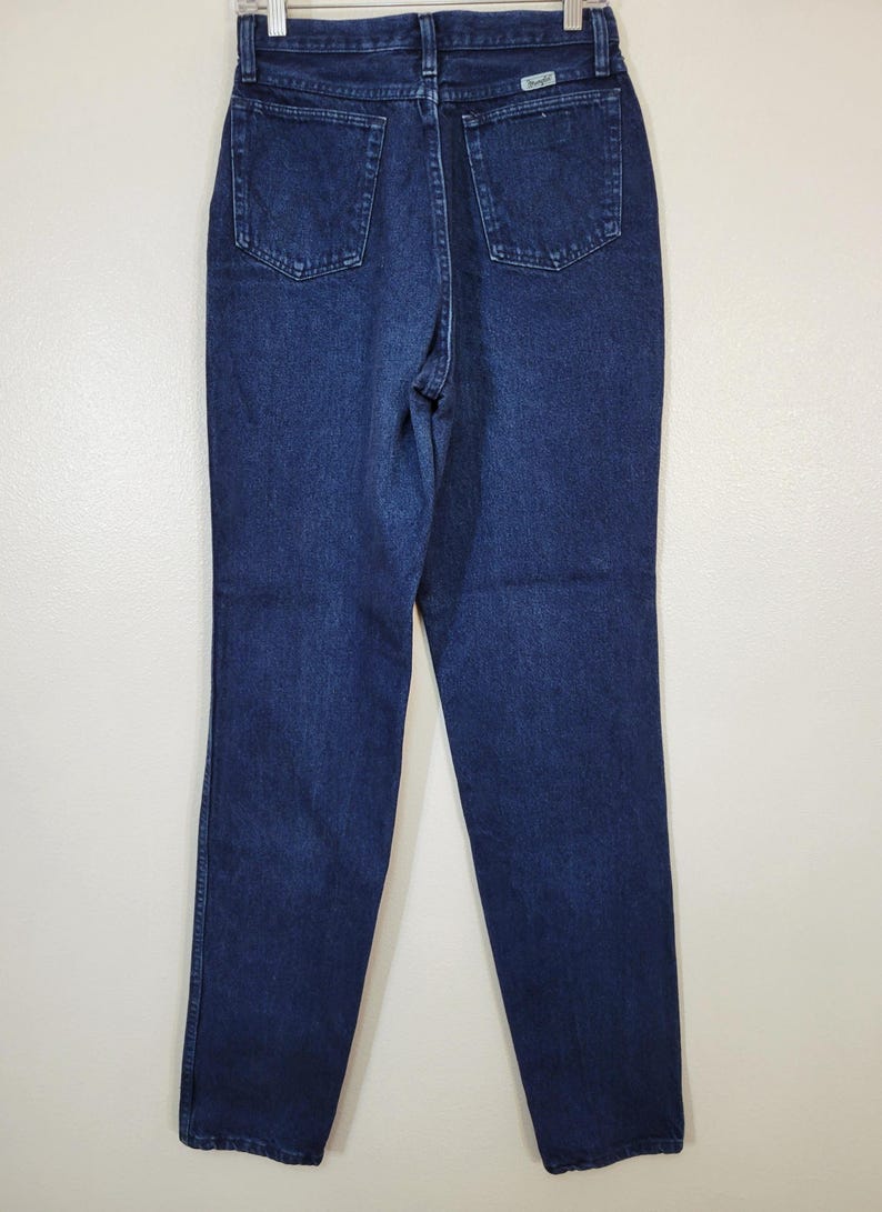 May include: Dark blue denim jeans with a classic five-pocket design. The jeans have a straight leg cut and a high waist. The Wrangler brand label is visible on the back pocket. The jeans are hanging on a hanger against a white background.