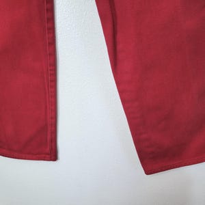 以下が含まれることがあります： 赤いデニムパンツのクローズアップ。パンツは無地の鮮やかな赤色です。画像は、脚と裾を含むパンツの下部を示しています。生地は中厚手のデニムのようです。