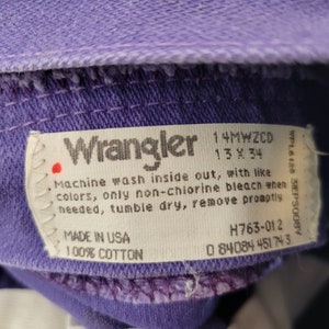 May include: Close-up of a purple Wrangler jeans label. The label includes the Wrangler logo, size 13 x 34, and washing instructions. Made in the USA from 100% cotton. The jeans are a vibrant purple color.