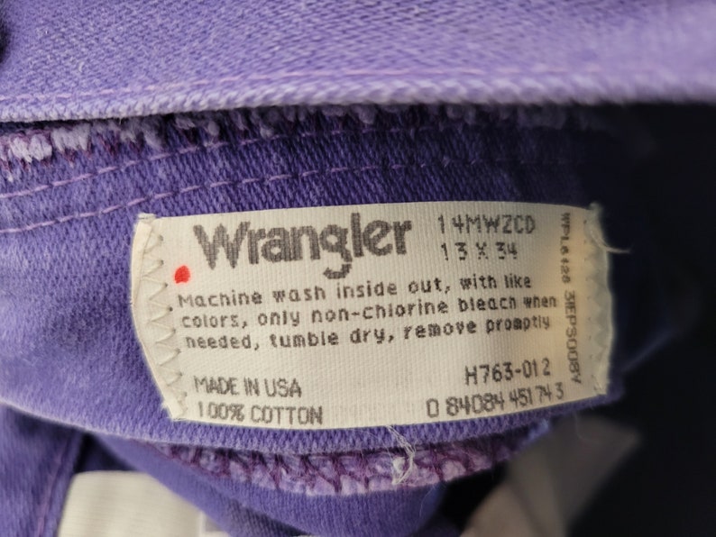 May include: Close-up of a purple Wrangler jeans label. The label includes the Wrangler logo, size 13 x 34, and washing instructions. Made in the USA from 100% cotton. The jeans are a vibrant purple color.