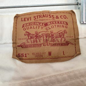 May include: Close-up of a brown Levi Strauss & Co. label on white denim. The label features text including "Original Riveted Quality Clothing" and "551™ Relaxed Fit Petite". The label has a graphic of two horse-drawn carriages.