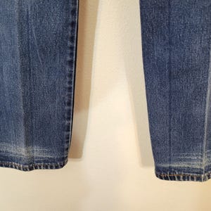 May include: Close-up of the lower portion of blue denim jeans. The jeans have a classic, straight-leg cut with a faded effect near the hem. The stitching is visible along the seams and hemline, showcasing the garment's construction.