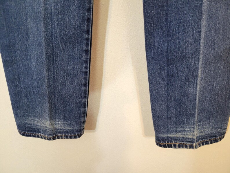 May include: Close-up of the lower portion of blue denim jeans. The jeans have a classic, straight-leg cut with a faded effect near the hem. The stitching is visible along the seams and hemline, showcasing the garment's construction.